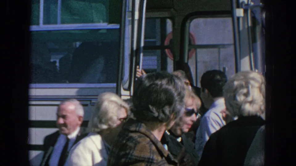 Older adults in coats and sunglasses pass beside a white bus in this 1966 Spain home-movie frame.