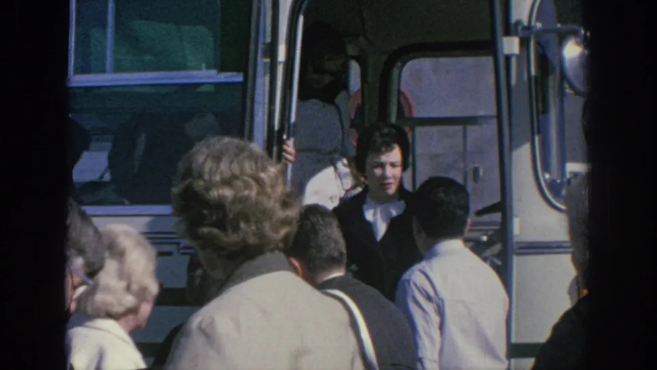 A group of adults stands around the open doorway of a white bus, with one younger-looking adult visible inside the entrance.