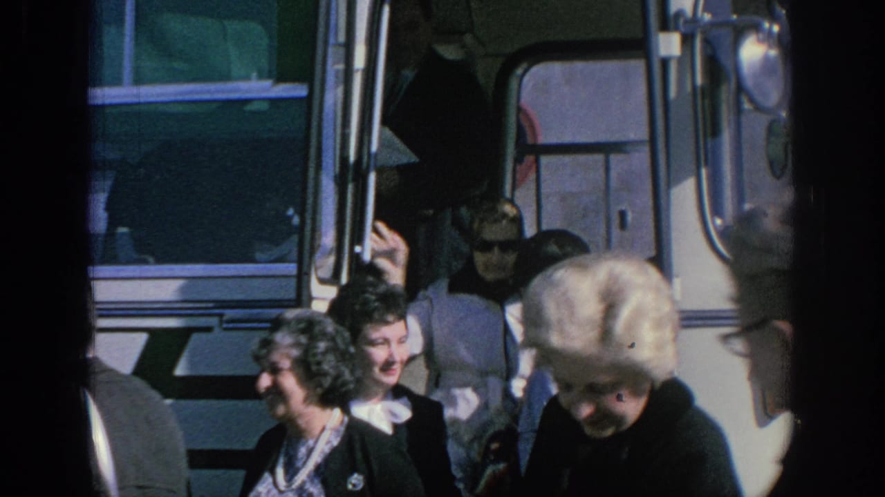 People stand near the side of a white bus, with faces and period hairstyles visible.
