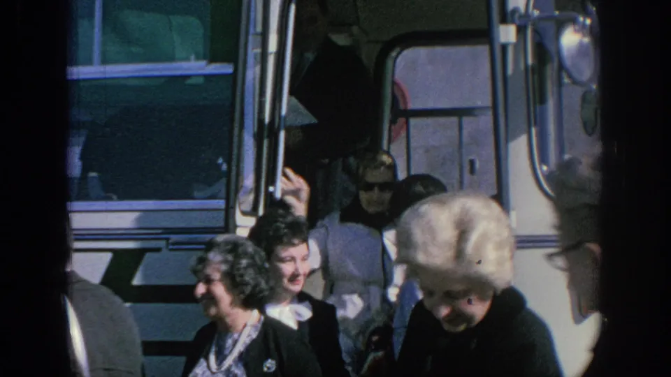 Adults in 1966 Spain stand beside a white bus, including women in dark coats and a blonde woman in the foreground.
