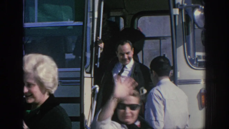 In Spain in 1966, a suited man exits a white bus as a woman in sunglasses waves close to the camera.