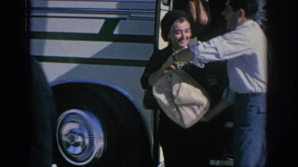 Beside a white bus in 1966 Spain, a man assists a woman holding luggage or folded fabric near the bus wheel.