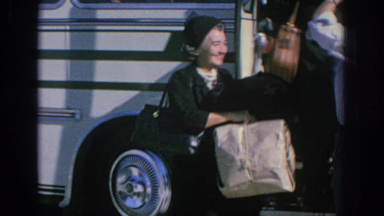 In Spain 1966 home-movie footage, an elderly woman in a dark coat and headscarf stands near a bus with bags in hand.