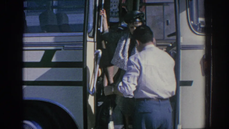 A woman in a dark coat descends from the bus doorway while a man in a light shirt stands close by to assist.