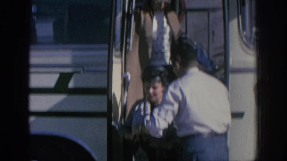 Near the bus door, an elderly woman in dark clothing is close to the ground while a man in a light shirt reaches toward her.