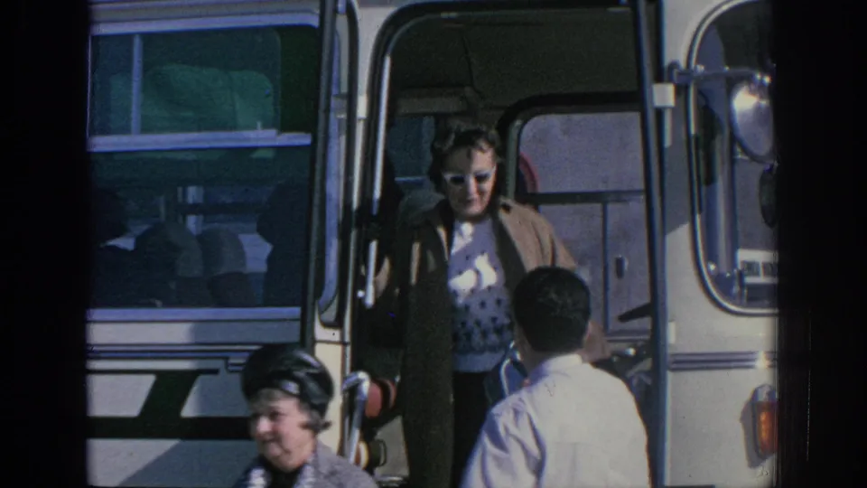 1966 Spain home-movie frame showing adults in sunglasses near the open doorway of a white and green tour bus.
