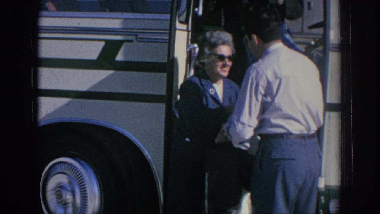A woman in sunglasses stands near a bus while a man faces her in the foreground.