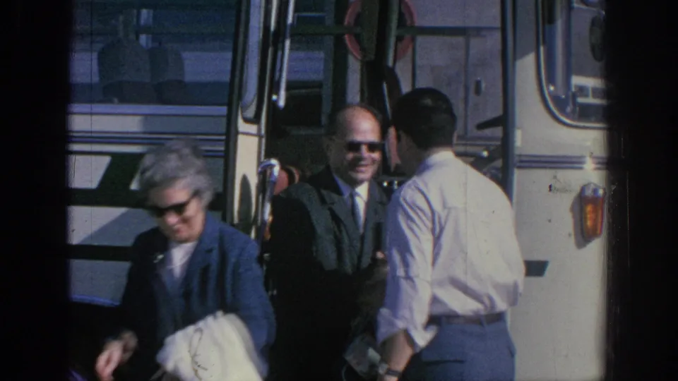 1966 Spain home-movie frame showing adults in mid-century travel clothes gathered beside a striped tour bus.