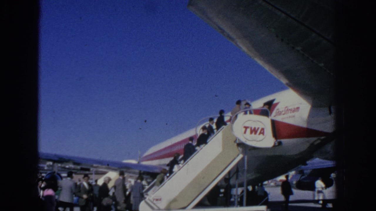1966 Spain home-movie frame showing passengers boarding a TWA aircraft by mobile stairs under a clear blue sky.