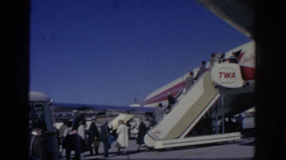 People in period clothing wait on the airport tarmac near mobile stairs and a TWA aircraft during a 1966 Spain travel scene.