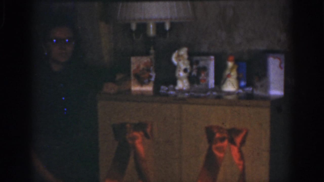 Shadowed woman stands beside a Christmas-decorated cabinet with bows, cards, ornaments, and a lamp.