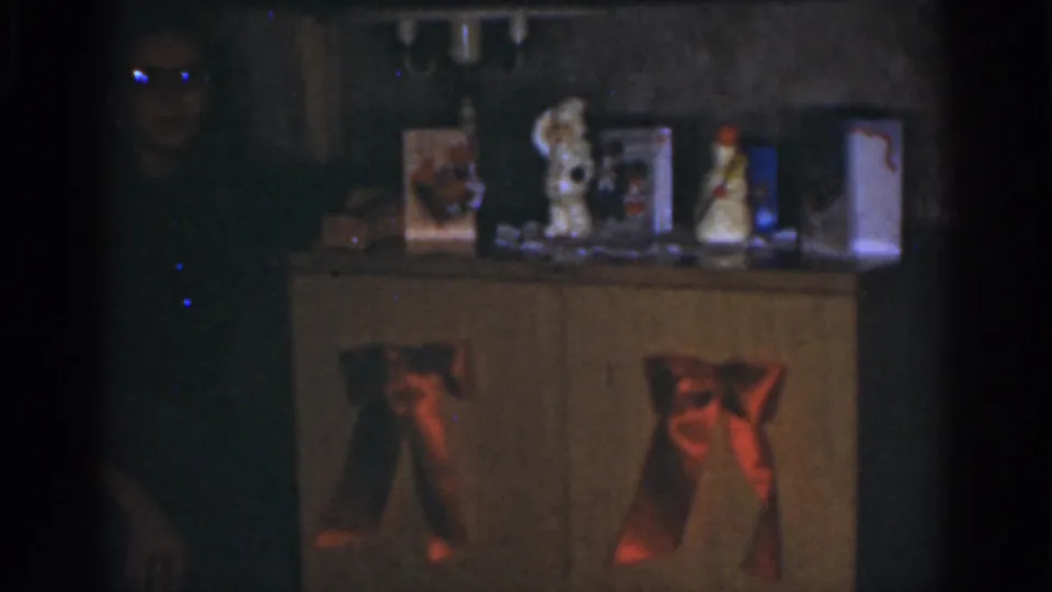 The frame shows a dim 1968 Omaha home scene with a woman near a decorated cabinet, red bows on the doors, and holiday objects on top.