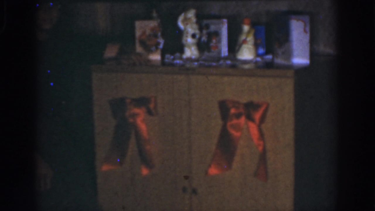 Dim interior with a wooden cabinet, red bows, tabletop ornaments, and a shadowed person at left.