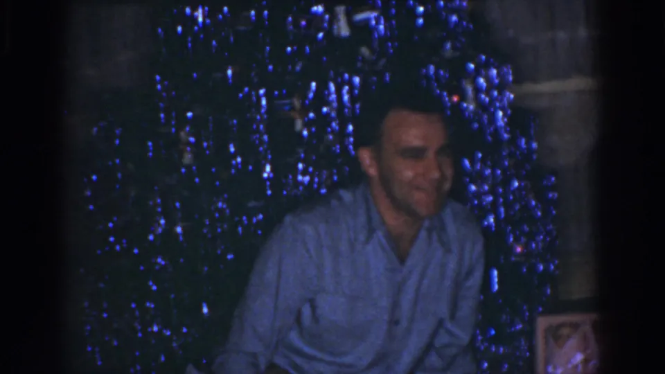 1968 Omaha home-movie frame showing a smiling man near a shimmering blue tinsel Christmas tree in a dim interior.