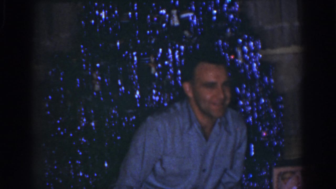 Adult man in Omaha 1968 home-movie footage sits before a dim Christmas tree glowing with blue lights.