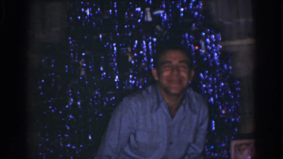 The man smiles toward the camera in 1968 Omaha home-movie footage, framed by blue Christmas lights.