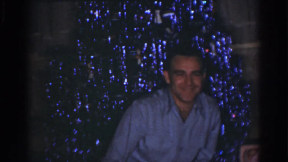 A seated adult man smiles in a dim Omaha holiday interior, with blue Christmas lights behind him.
