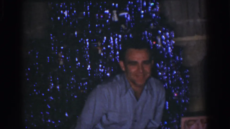 The man faces the camera with a slight smile as the blue-lit Christmas tree fills the 1968 Omaha home-movie background.