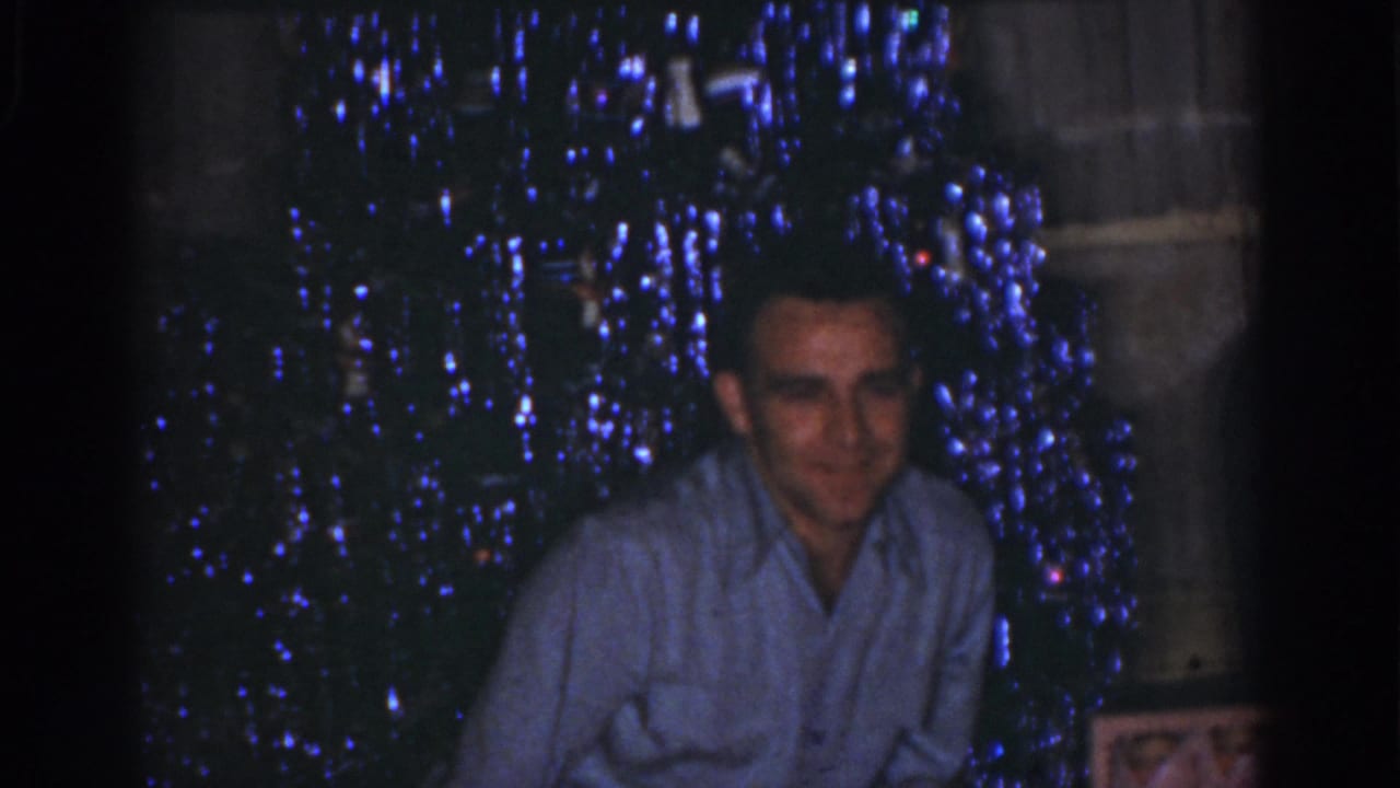 1968 Omaha home-movie frame of a man smiling toward the camera beside a dark Christmas tree lit with blue lights.
