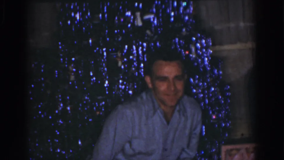 1968 Omaha home-movie frame of a man smiling toward the camera beside a dark Christmas tree lit with blue lights.