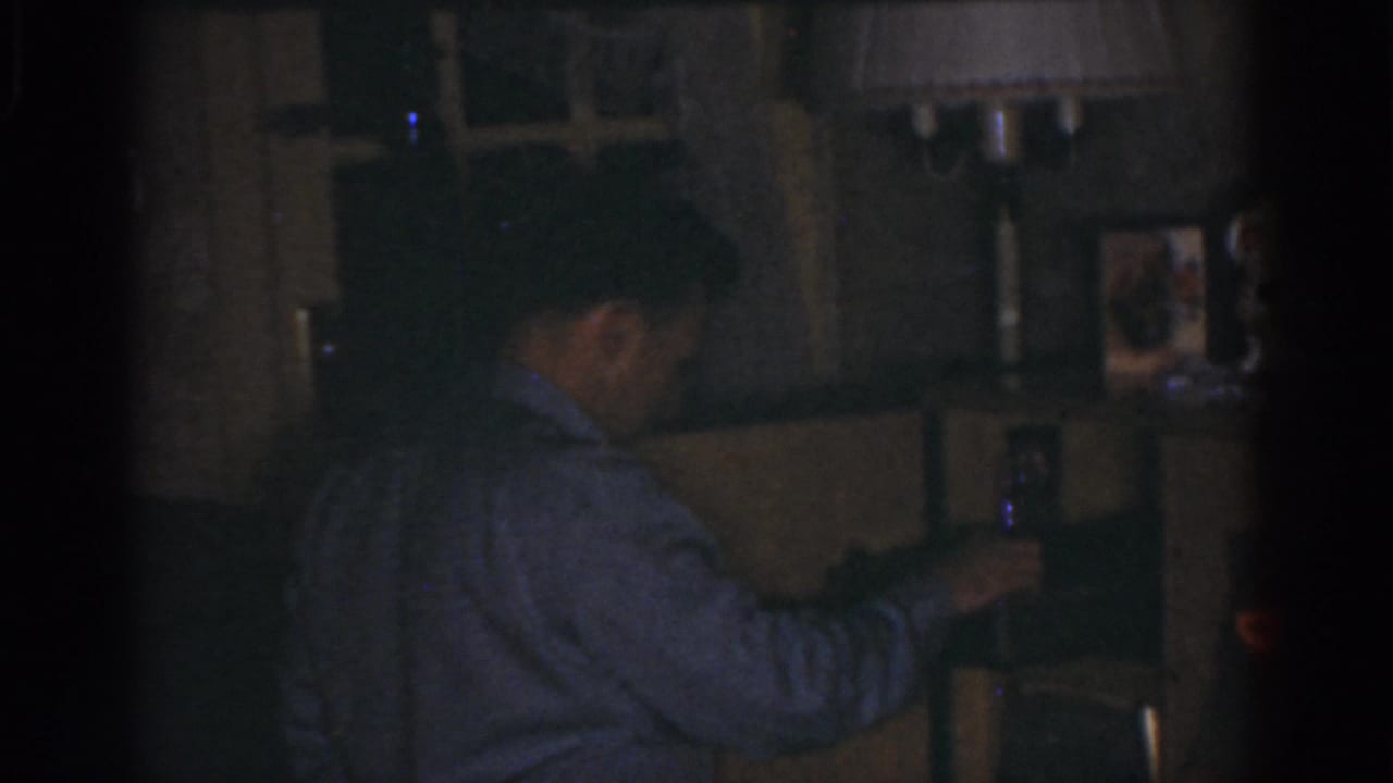 Man turned sideways reaching toward items on a shelf or cabinet in a dim room.