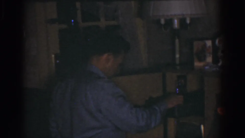 1968 Omaha home-movie frame of the man turned from camera, reaching toward household items on shelving or cabinetry.