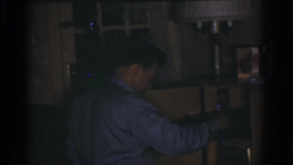 1968 Omaha home-movie frame showing the man in profile as he handles an object near a dark shelf or cabinet.