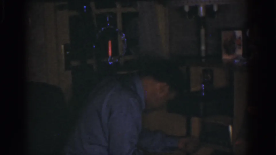 1968 Omaha home-movie frame of the man bending near shelves and household items, with blue light reflections in the dark room.