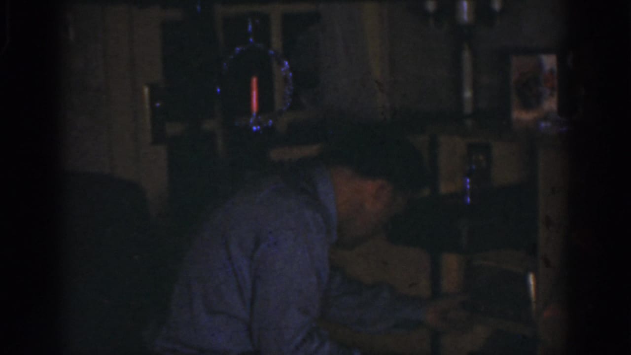 In 1968 Omaha home-movie footage, an adult man bends toward a tabletop device in a dark private interior.