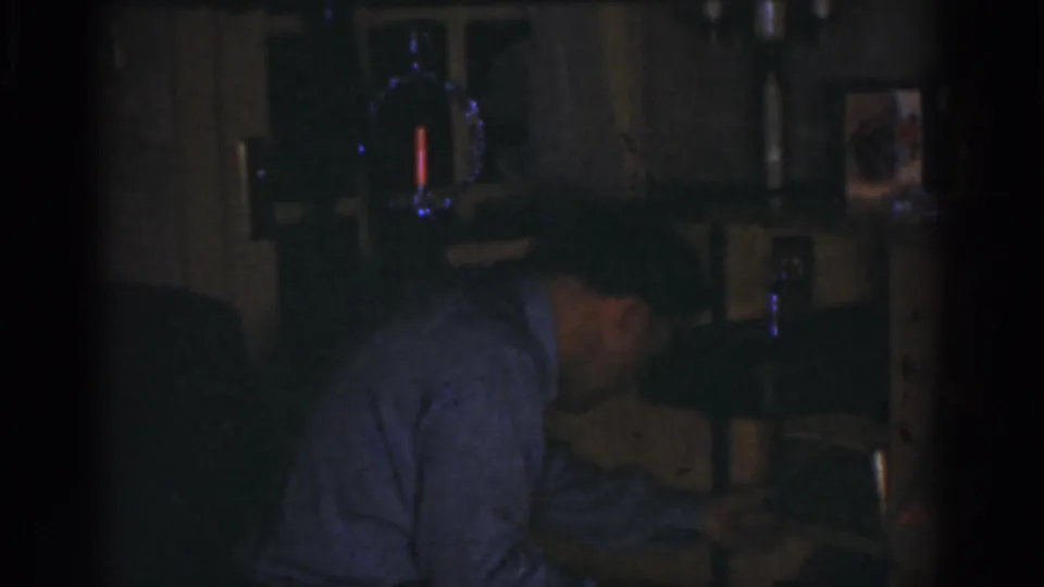 In 1968 Omaha home-movie footage, an adult man bends toward a tabletop device in a dark private interior.