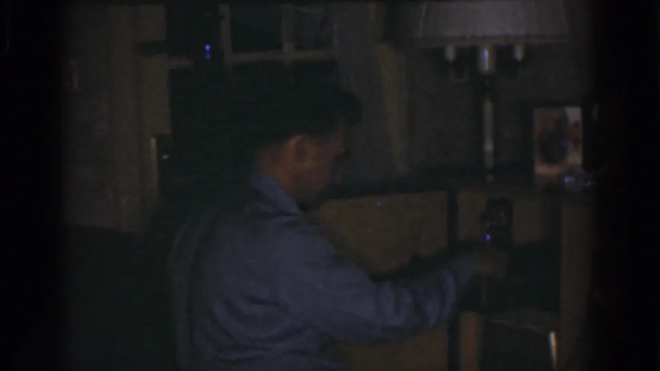 A 1968 home-movie frame shows the man looking down at equipment near a small red indicator light in a private room.