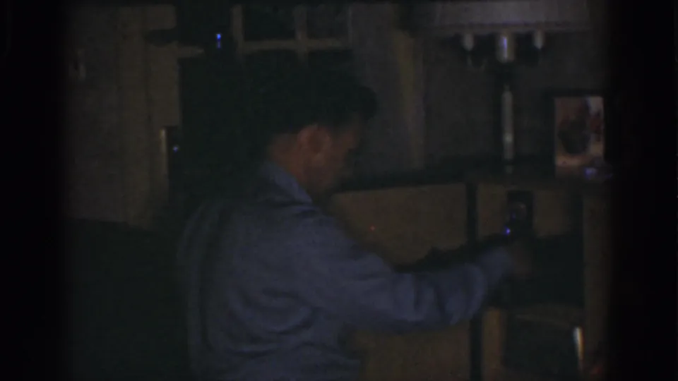 The man appears to adjust or hold a small item near the device, with household pictures and furniture behind him.
