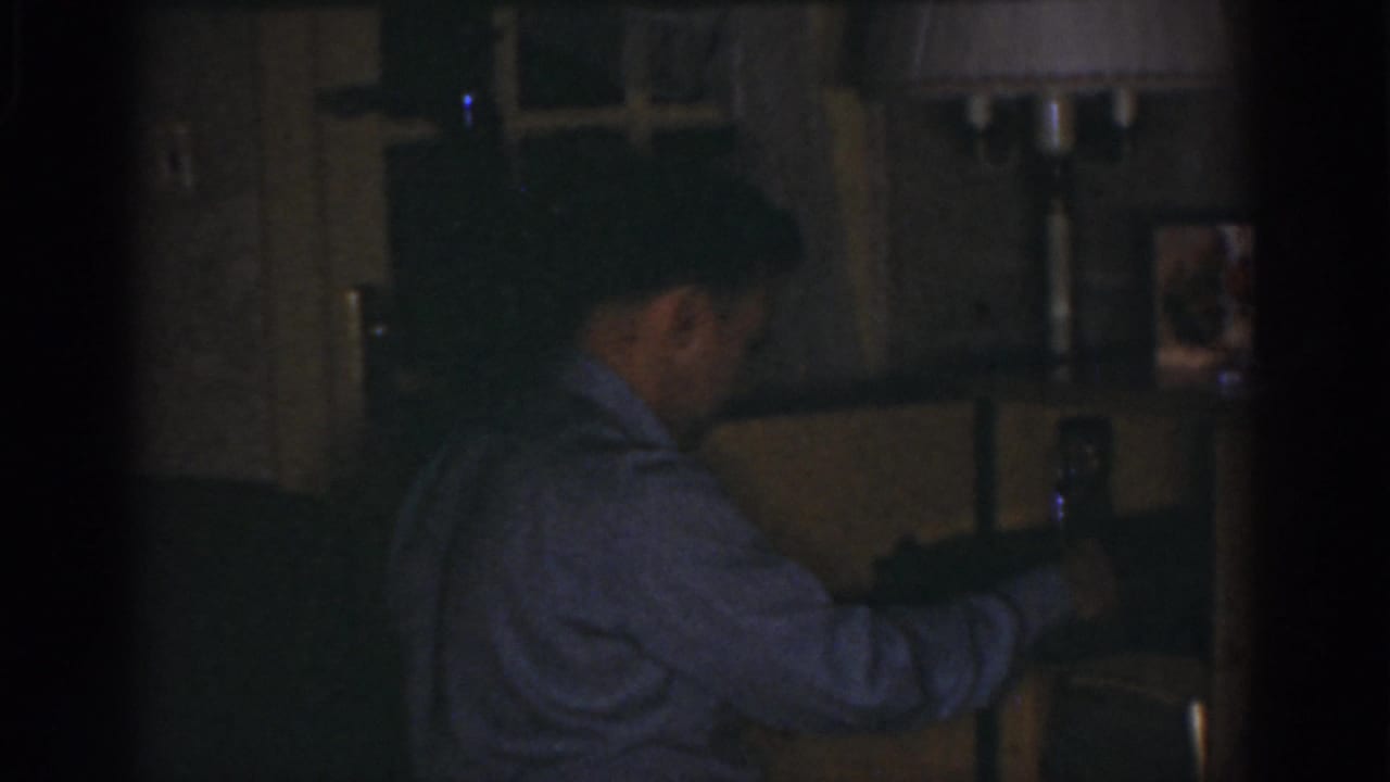In a dim Omaha home interior from 1968 metadata, an adult man in a light shirt reaches toward shelving near framed pictures.