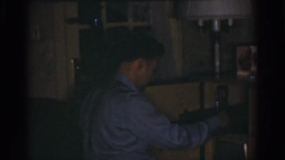 In a dim Omaha home interior from 1968 metadata, an adult man in a light shirt reaches toward shelving near framed pictures.