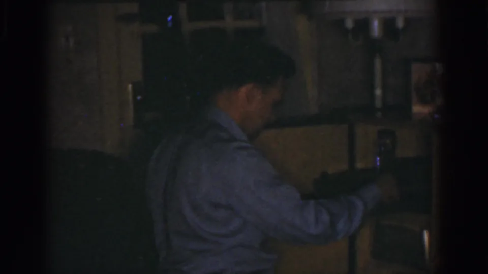 The man is seen in profile in the dim room, extending one hand toward a shelf with a small object nearby.