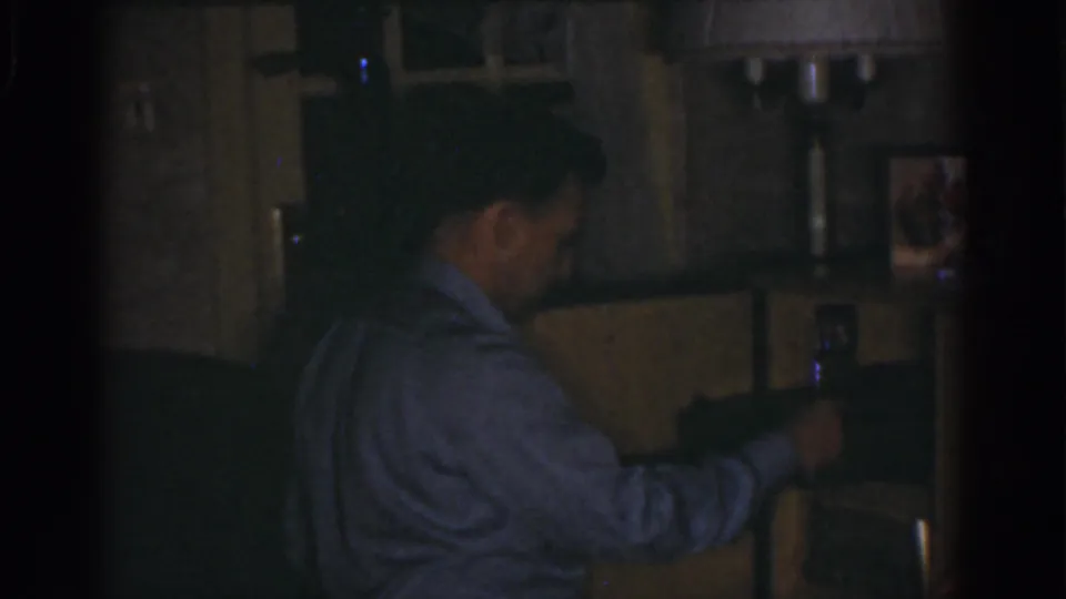 In the low-light home-movie frame, the man continues reaching toward a shelf area with framed pictures behind him.