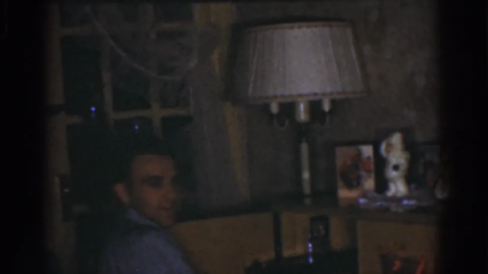 The man turns back toward the camera and appears to smile beside a table lamp, shelf, and framed photos in a private interior.