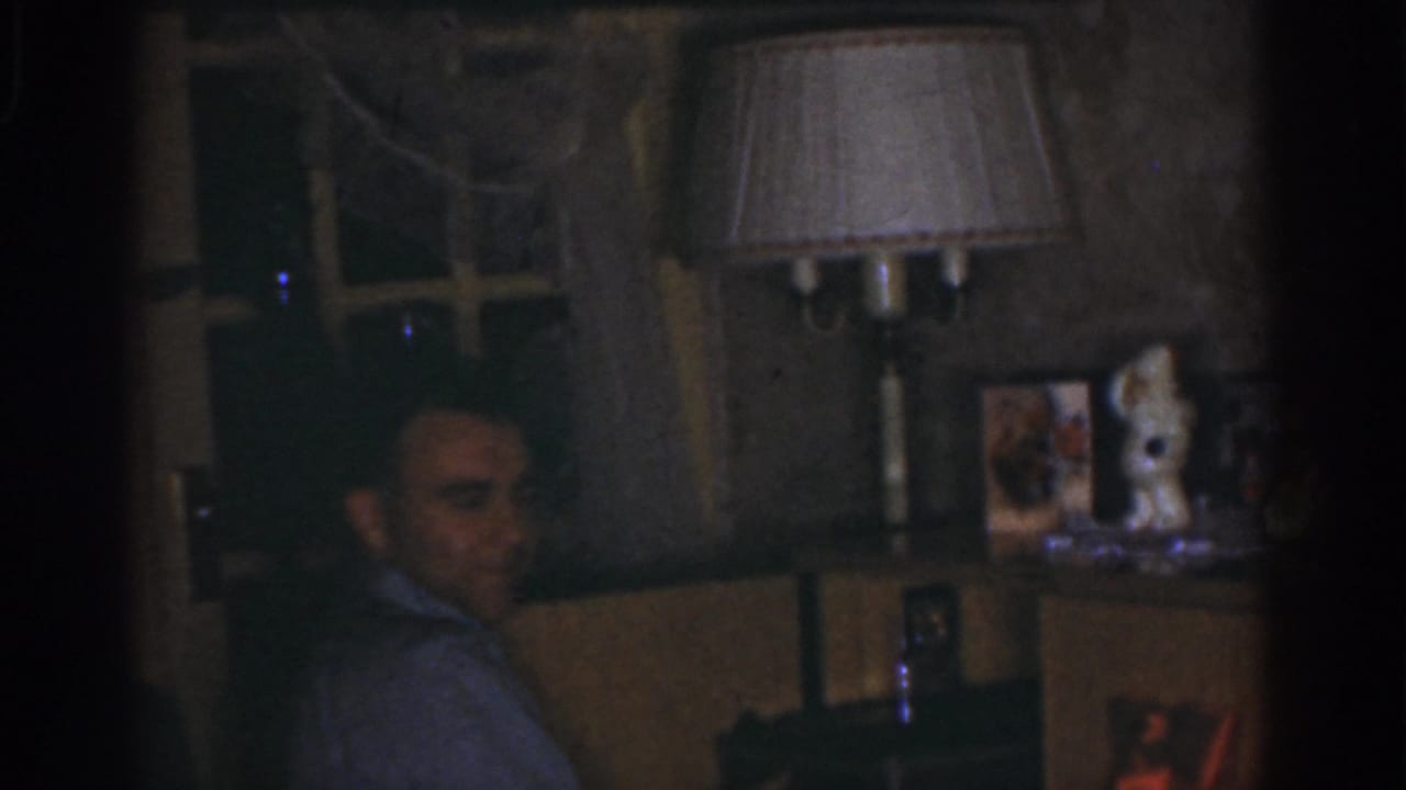 1968 Omaha home-movie frame showing a seated man in a dim private room with a lamp, window, framed photos, and figurines behind him.