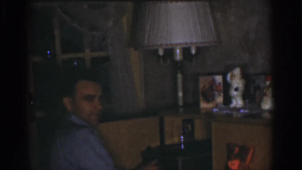 1968 Omaha interior frame with a seated adult man looking back toward the camera, lit by a table lamp in a private room.