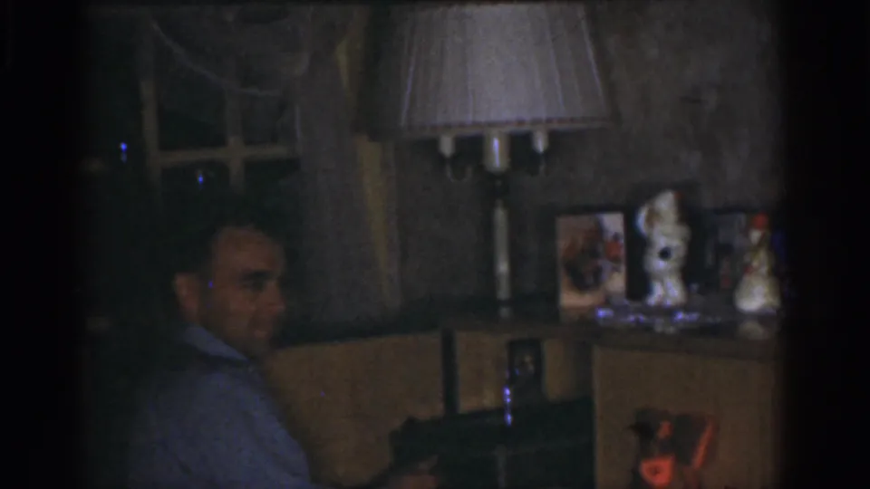 1968 Omaha home-movie still showing the man seated in profile in a low-lit room with domestic furnishings behind him.