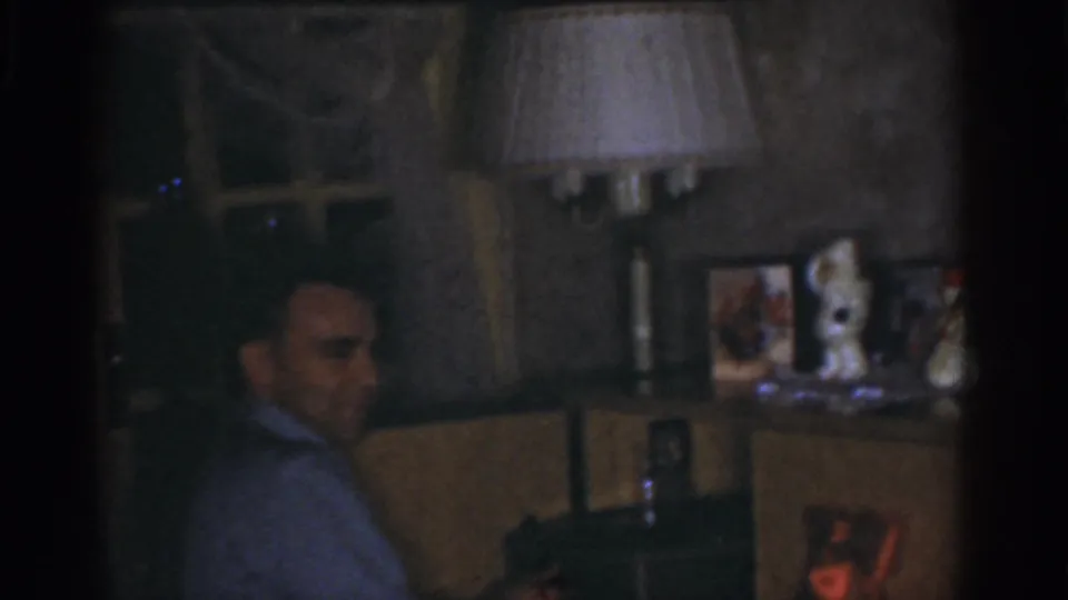 1968 Omaha home-movie frame of a seated man in partial profile, surrounded by a lamp, window, photographs, and small decorations.