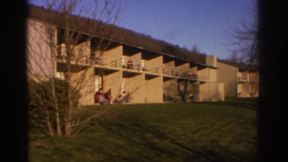 A warm 8mm exterior view shows the lodge building, balcony railings, trees, lawn shadows, and people gathered on the grass.