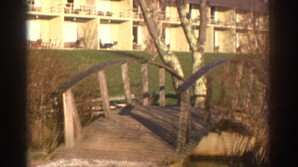 1971 Bedford, Virginia 8mm frame showing a small wooden bridge over a stream area, with a balcony building and bare tree behind it.