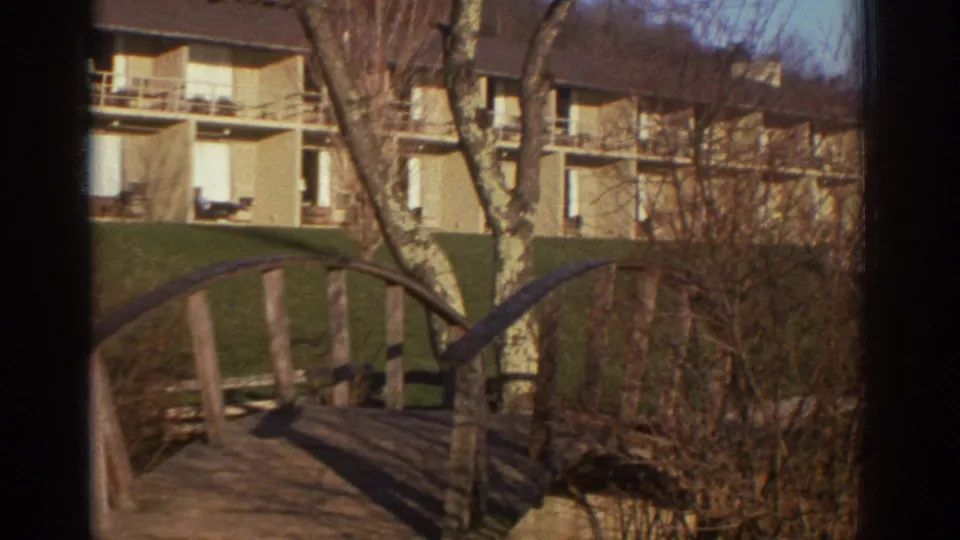 1971 Bedford, Virginia 8mm frame of the wooden bridge, shrubs, lawn, bare trees, and low balcony building on the property grounds.