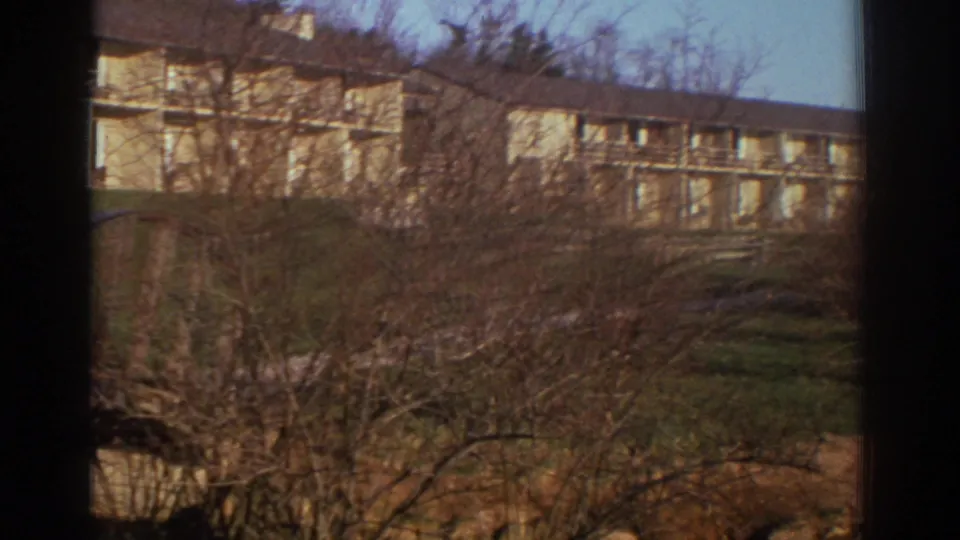 A 1971 home-movie view of a Bedford apartment block behind leafless shrubs and winter-bare trees.