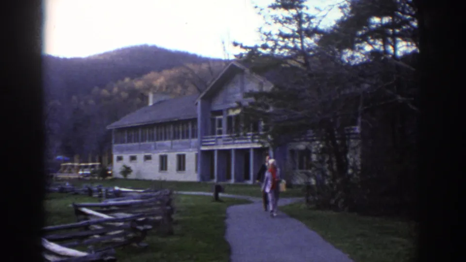 1971 home-movie frame of an older couple walking together near a split-rail fence and lodge-style building in a mountain valley.