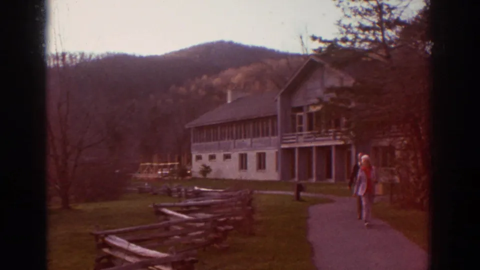 Older couple walks along a curving path past a split-rail fence, with a lodge-style building and wooded mountain behind them.