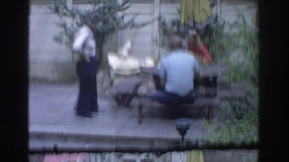 1971 Miami home-movie frame showing adults on a backyard patio with a picnic table, shrubs, fence, and white garden fountain.