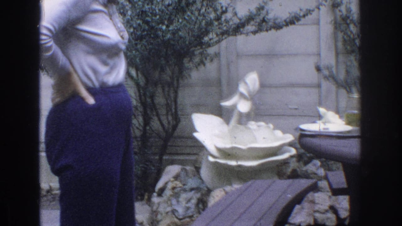 Woman in flared pants stands beside shrubs and a white garden fountain.