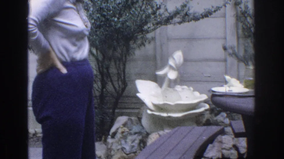1971 Miami backyard frame with a woman in a light top and dark flared pants near shrubs, rocks, and a white ceramic fountain.